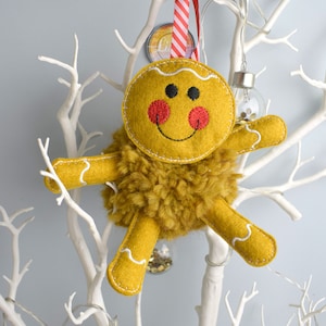 May include: A gingerbread man Christmas ornament made of felt with a fuzzy brown body and yellow icing details. The ornament is hanging from a white branch with red and white striped ribbon.