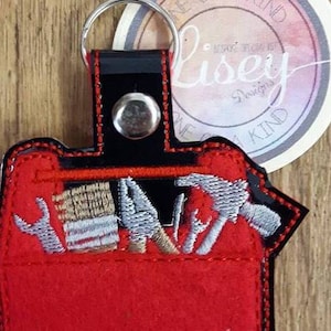 May include: Red felt toolbox keychain with embroidered tools, including a hammer, wrench, and screwdriver. The keychain has a black leather strap and a silver metal ring. The text "One of a Kind" is visible on a white circle behind the keychain.