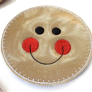 May include: A round, beige, glittery coaster with a white stitched border. The coaster has an embroidered smiley face with black eyes, a black smile, and red cheeks.