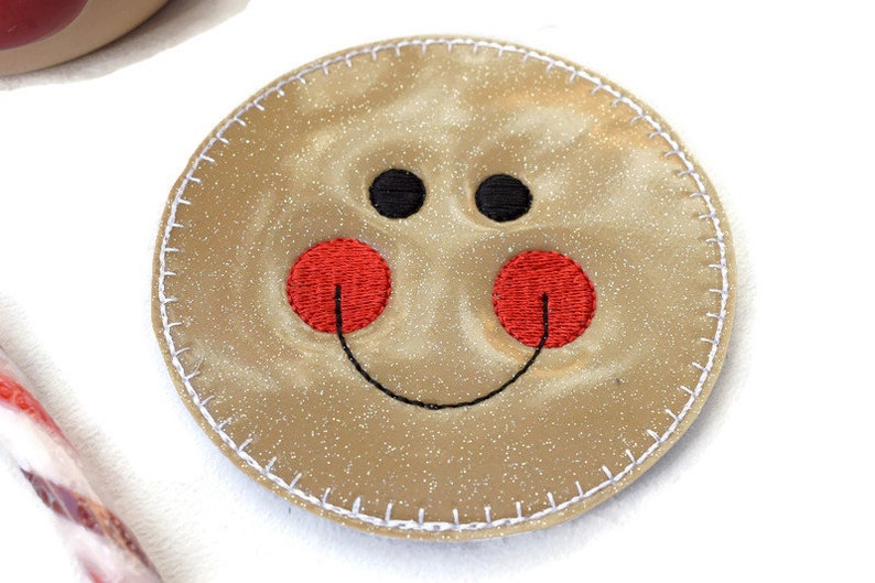 May include: A round, beige, glittery coaster with a white stitched border. The coaster has an embroidered smiley face with black eyes, a black smile, and red cheeks.