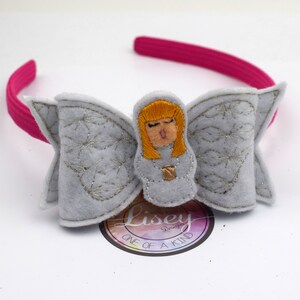 May include: A grey felt headband with a silver glitter bow and a felt angel with blonde hair and a gold halo. The headband is pink. The bow has a silver glitter design. The angel is wearing a white dress with a gold halo. The headband says "Lisey Designs One of a Kind".