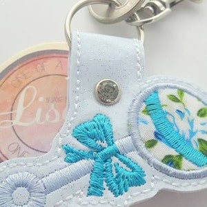 以下が含まれることがあります： 青い刺繍のガラガラ模様が施された白い布製のキーホルダー。キーホルダーには銀色のリングと銀色の留め具が付いています。ガラガラには青いリボンと透明なラインストーンが付いています。