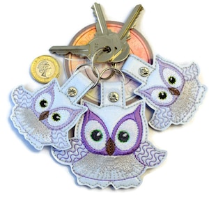 May include: Three purple and white embroidered owl keychains with a silver key ring. The owls have large eyes and a brown beak.
