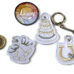 Könnte beinhalten: Vier weiße Stoffschlüsselanhänger mit goldener Stickerei. Die Schlüsselanhänger zeigen eine Hochzeitstorte, zwei Glocken und ein Hufeisen mit einem Herz. Ein silberner Schlüsselring ist im Lieferumfang enthalten.