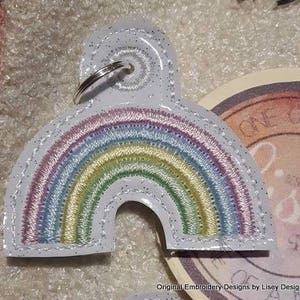 以下が含まれることがあります： 銀色のキーリングが付いた白い虹のキーホルダー。虹はパステルピンク、ブルー、グリーン、イエローの糸で刺繍されています。