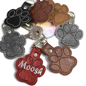 May include: A set of six leather paw print keychains. The keychains are in various colours, including black, brown, grey, and red. One keychain has the name "Moosh" embroidered on it.