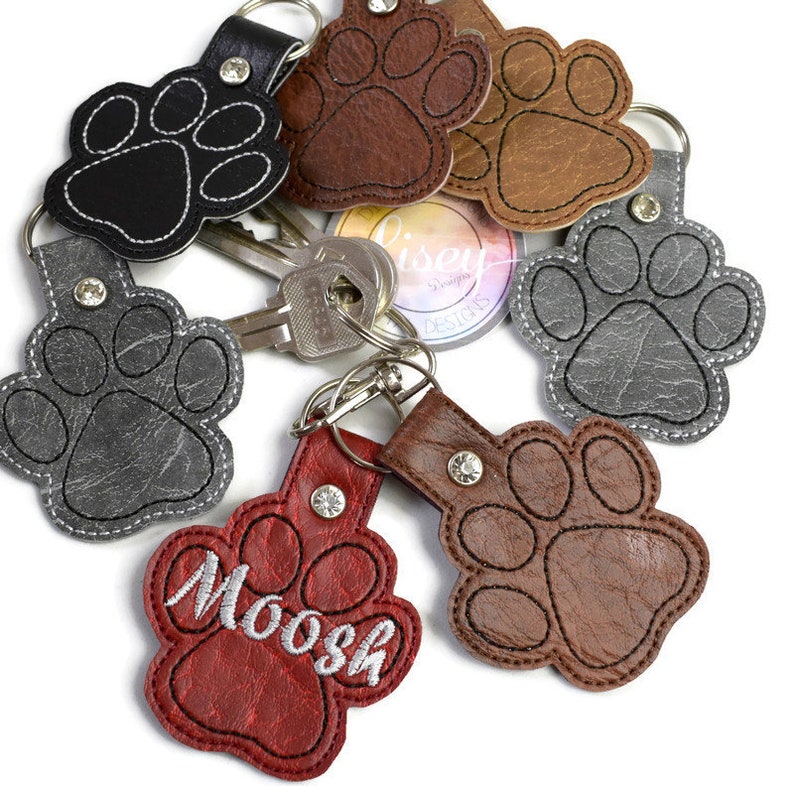 May include: A set of six leather paw print keychains. The keychains are in various colours, including black, brown, grey, and red. One keychain has the name "Moosh" embroidered on it.