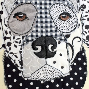 May include: A black and white patchwork fabric dog with a black nose and brown eyes. The dog is stitched onto a cream-colored background.