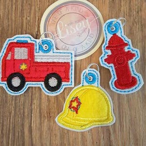 May include: Three embroidered patches featuring a red fire engine, a yellow firefighter helmet, and a red fire hydrant. The patches are all outlined in blue and have a white star in the centre.