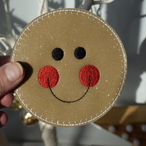May include: A round, brown, glittery ornament with a white stitched border. The ornament has a smiling face with black eyes and a red embroidered mouth.