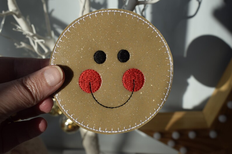 May include: A round, brown, glittery ornament with a white stitched border. The ornament has a smiling face with black eyes and a red embroidered mouth.