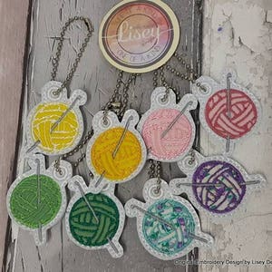 May include: Seven colorful embroidered keychains shaped like balls of yarn with knitting needles. Each keychain has a different color yarn and a white background. The keychains are labeled "Lisey One of a Kind".