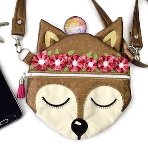 Könnte beinhalten: Eine kleine braune und weiße Umhängetasche in Form eines Fuchses mit Reißverschluss. Die Tasche hat einen rosa Quasten-Reißverschluss und ein florales gesticktes Stirnband mit roten und weißen Blumen. Die Tasche ist aus einem strukturierten Stoff gefertigt und hat einen braunen Riemen.