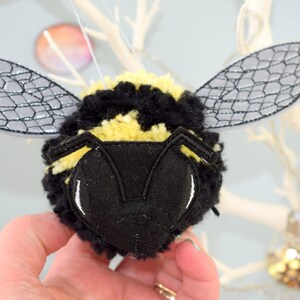 May include: A black and yellow bumble bee pom-pom ornament with gray felt wings. The bee has a black felt face with white eyes.