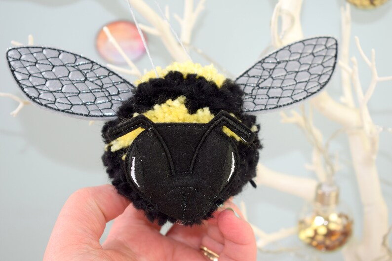 May include: A black and yellow bumble bee pom-pom ornament with gray felt wings. The bee has a black felt face with white eyes.