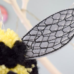 May include: A close-up of a gray felt bee wing with a black embroidered honeycomb pattern. The wing is attached to a yellow and black pom-pom bee.