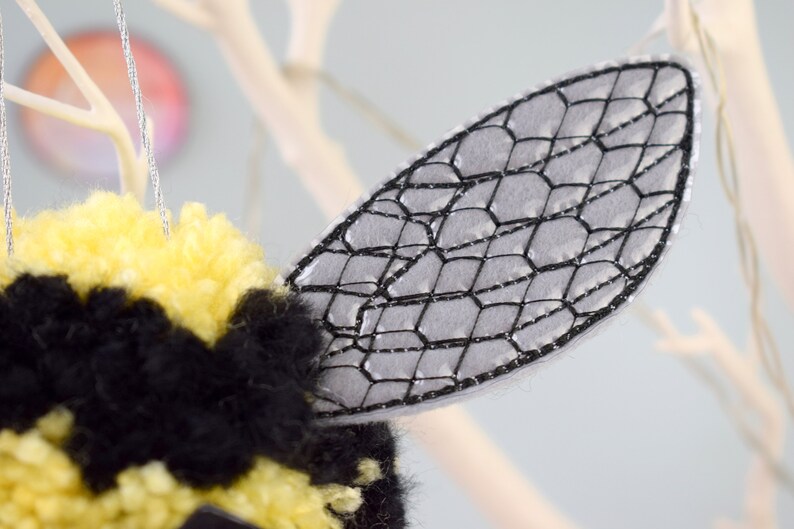 May include: A close-up of a gray felt bee wing with a black embroidered honeycomb pattern. The wing is attached to a yellow and black pom-pom bee.