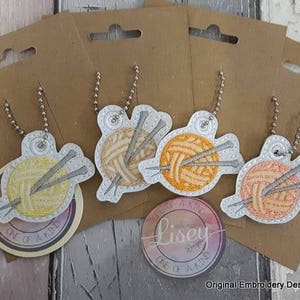 May include: Five colorful embroidered keychains featuring a ball of yarn and knitting needles. Each keychain is attached to a silver chain and has a white background with the text "One of a Kind" printed on it.