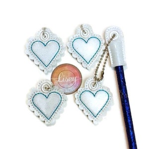May include: Four white heart-shaped charms with a teal outline and a sparkly finish. Each charm has a small silver loop at the top. The charms are attached to a gold chain and a blue pen with a white cap.