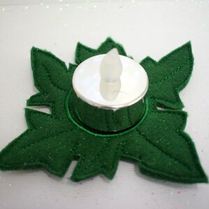 Może przedstawiać: Zielony świecznik z filcu w kształcie płatka śniegu z centralnie umieszczonym srebrnym świecznikiem na tealight.