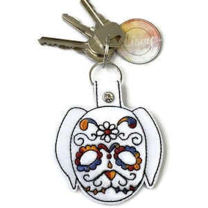 Peut inclure: Porte-clés en feutre blanc avec un motif de chien en sucre brodé. Le crâne est décoré de fleurs colorées et de volutes. Le porte-clés est attaché à un anneau de clé argenté.