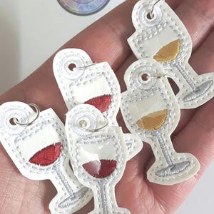 May include: Five white felt wine glass charms with silver stitching. Each charm has a different coloured liquid inside: red, gold, and clear. The charms have a silver loop for attaching to a glass.