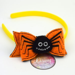 May include: A yellow headband with an orange and black felt spider bow. The spider has a stitched-on face with two eyes and a stitched-on mouth. The bow is attached to the headband with a knot. The headband is a one-of-a-kind design by Lisey Designs.