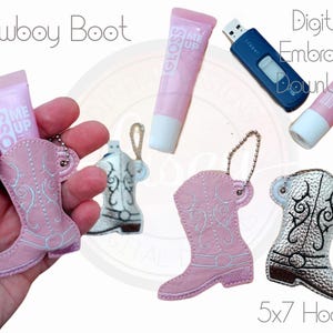 Może przedstawiać: Cyfrowy wzór haftu przedstawiający kowbojskie buty w kolorze różowym i srebrnym. Wzór jest pokazany na różowym błyszczyku do ust, srebrnym breloczku i pendrive'ie. Wzór jest przeznaczony do obręczy 5x7.