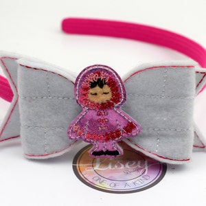 May include: A pink headband with a gray felt bow and a pink felt girl wearing a pink coat and a red hood. The girl is embroidered with details and has a white face. The headband is decorated with white stitching.