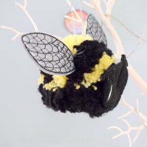 May include: A black and yellow yarn bee ornament with embroidered wings. The bee is hanging from a white branch.
