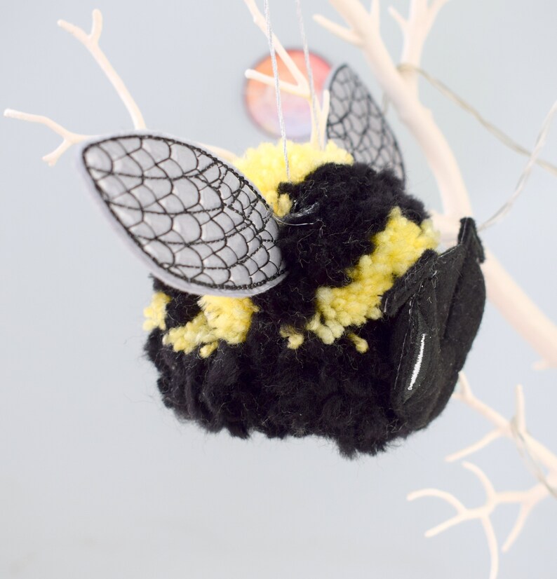 May include: A black and yellow yarn bee ornament with embroidered wings. The bee is hanging from a white branch.