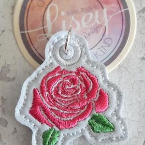 May include: Embroidered rose patch featuring a red flower and green leaves on a white background. The patch has a silver border and a metal ring. A round tag with text is in the background.