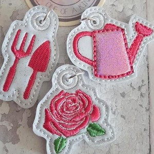 May include: Three white felt ornaments with pink embroidery. The ornaments feature a fork and knife, a watering can, and a rose.