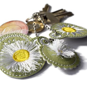 May include: A green leather keychain with three embroidered white daisies with yellow centres. The keychain also has a silver key ring with keys attached.
