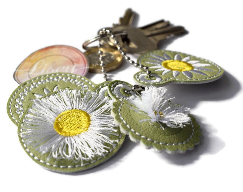 May include: A green leather keychain with three embroidered white daisies with yellow centres. The keychain also has a silver key ring with keys attached.