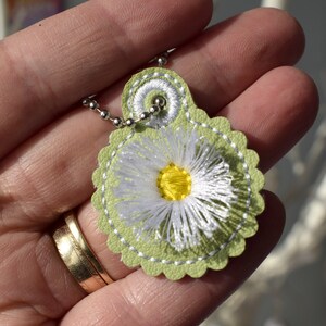 May include: A green leather pendant with a white daisy embroidered on it. The pendant is attached to a silver chain.
