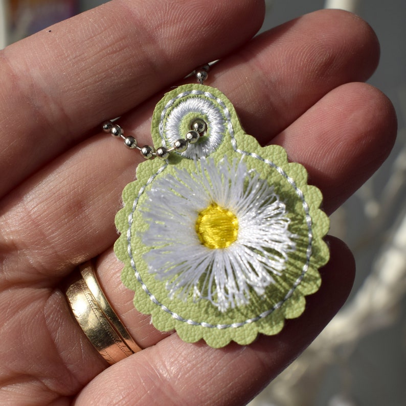May include: A green leather pendant with a white daisy embroidered on it. The pendant is attached to a silver chain.