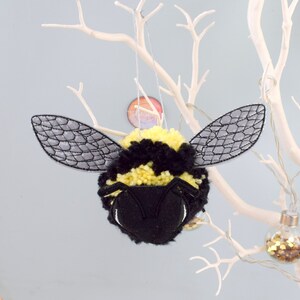 May include: A black and yellow pom-pom bee ornament with gray felt wings. The bee has a black face with white eyes and a black and yellow striped body.