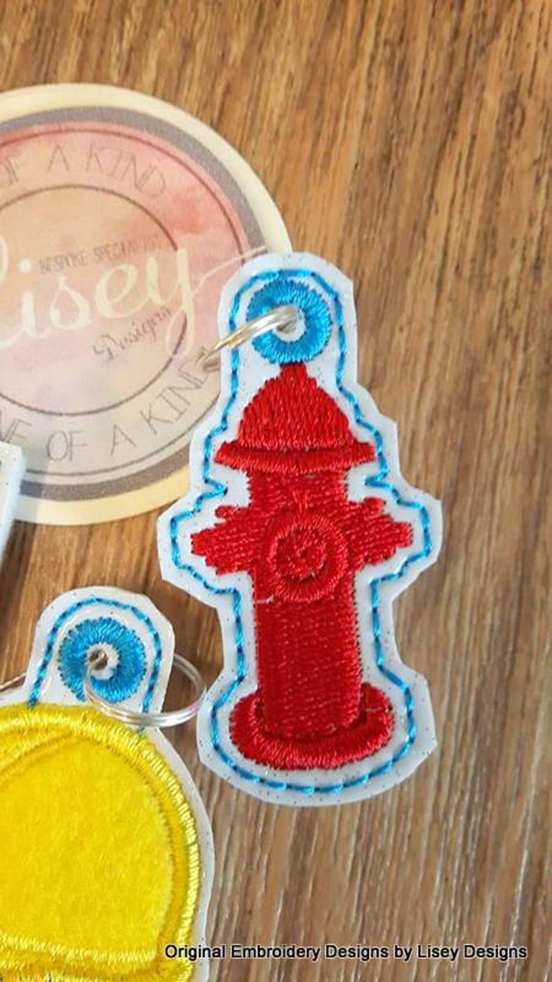 May include: A red and white embroidered fire hydrant keyring with a blue ring. The keyring is made of felt and has a white outline.