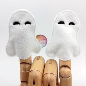 以下が含まれることがあります： 黒い刺繍の目をつけた白いフェルト製の幽霊指人形2つ。人形は木の手の上にある。