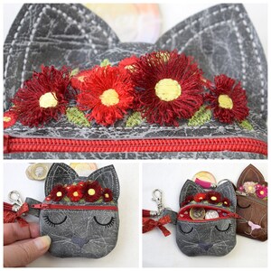 Pu&ograve; includere: Un portamonete grigio a forma di gatto con cerniera rossa e design floreale. La borsa presenta fiori rossi ricamati con centri gialli e foglie verdi. Il volto del gatto ha gli occhi chiusi e un piccolo naso.