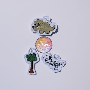 May include: Three felt patches featuring dinosaur and tree designs. The patches are brown, green and white. The patches have a loop for attaching to a bag or other item. The patches are labelled "Lisey Digital Designs".