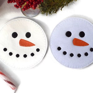 Peut inclure: Deux dessous-de-verre en feutre blanc en forme de bonhomme de neige avec des yeux noirs, une bouche noire et un nez orange. Les dessous-de-verre sont ronds et ont une bordure blanche cousue.