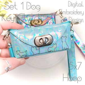 May include: A turquoise iridescent key clutch with a silver clasp and a silver charm. The clutch has a design of five dogs embroidered on it. The text "Set 1 Dog Key Clutch" and "Digital Embroidery Design 5x7 Hoop" is visible in the image.