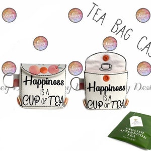 May include: White tea bag case with the text "Happiness is a cup of tea." One case features peach illustrations, the other a teacup. Includes a green tea bag. The cases have orange button closures and silver key rings.
