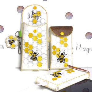 Puede incluir: Tres fundas de teléfono blancas con diseños de abejas y panales bordados. La funda más grande tiene el texto "Bee Happy" bordado en la parte superior. La funda más pequeña tiene una solapa de cuero marrón.