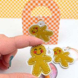 May include: Two felt gingerbread man ornaments with smiling faces and red cheeks. One ornament is attached to a silver key chain and the other is attached to a pencil.