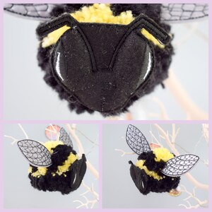 May include: A black and yellow bumble bee pom pom ornament with embroidered wings and eyes. The ornament is hanging from a branch.