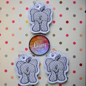 Puede incluir: Tres adornos de fieltro blanco con elefantes grises bordados. Cada adorno tiene un lazo plateado para colgar. Los adornos están sobre un fondo blanco con lunares rojos, marrones y verdes.