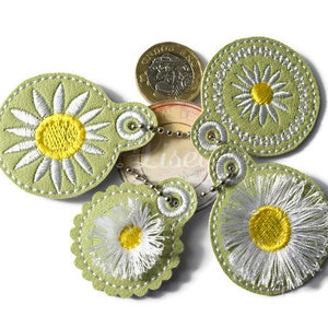 May include: Five green felt keyrings with daisy embroidery. Each keyring has a different style of daisy, with white petals and a yellow centre. The keyrings are connected by a silver chain.